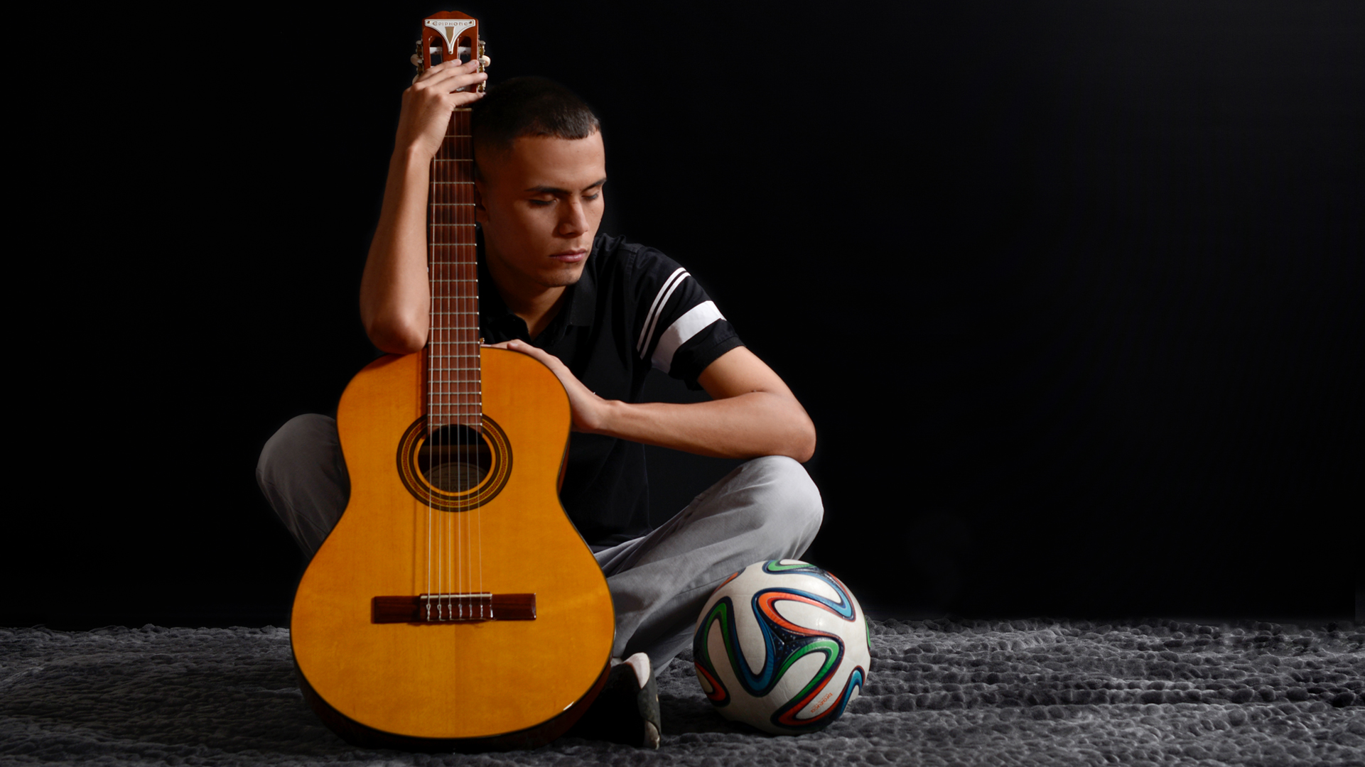 En la imagen: José Quiceno sostiene su guitarra sentado en el suelo junto a un balón de fútbol